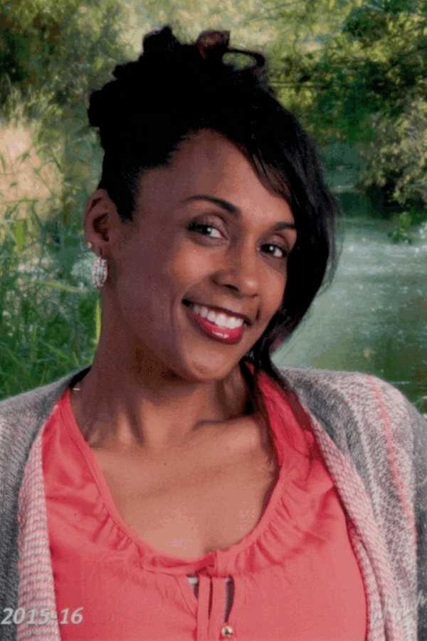 Woman smiles, wearing coral top and gray cardigan. Updo hairstyle, background of a stream.