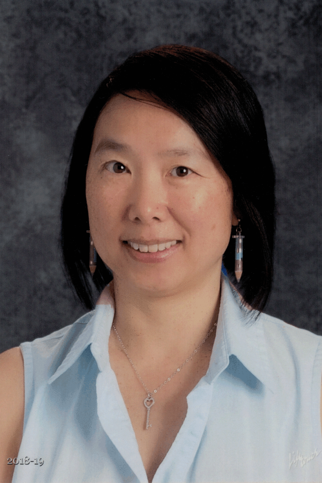 Woman with black hair, wearing light blue shirt, smiling, key necklace, earrings. Background: gray.