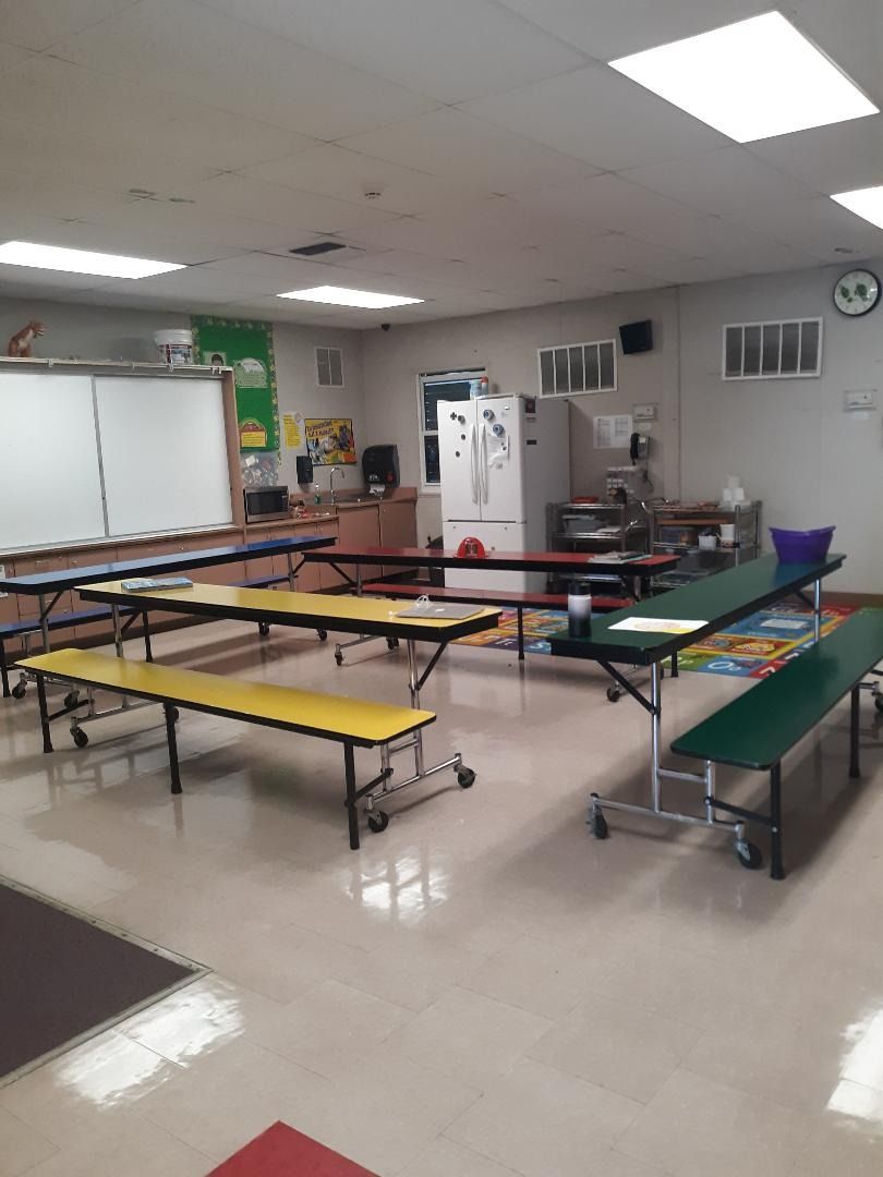 School cafeteria with long, colorful tables. Whiteboard, refrigerator, and microwave visible.