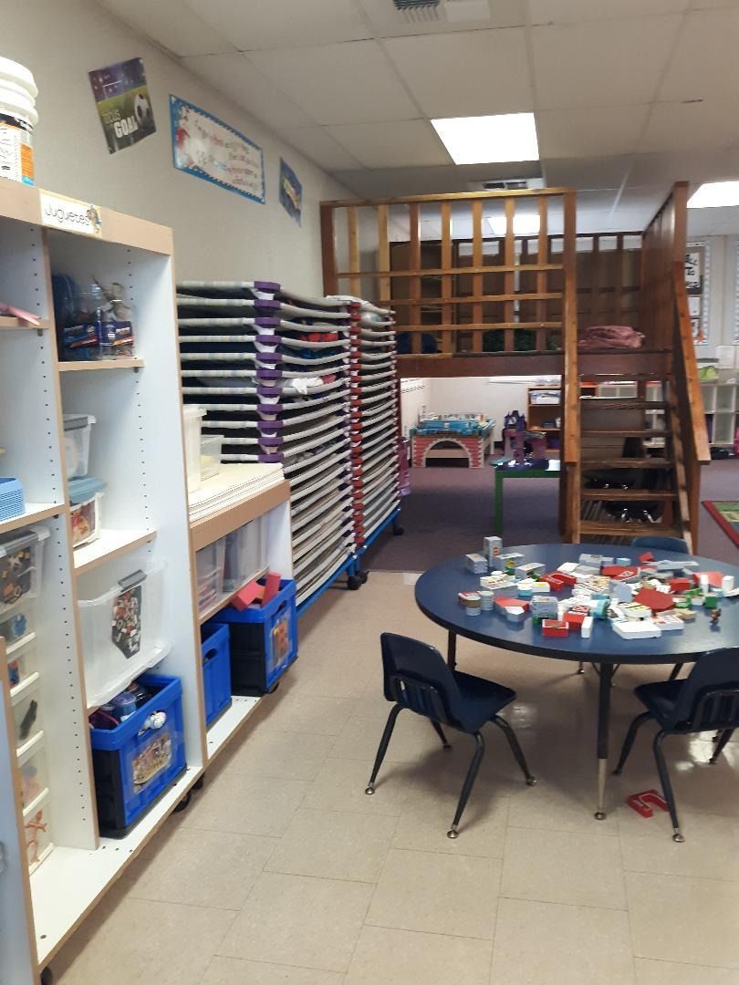 Classroom interior: shelving unit, art supply storage, round table with chairs, and a play loft.