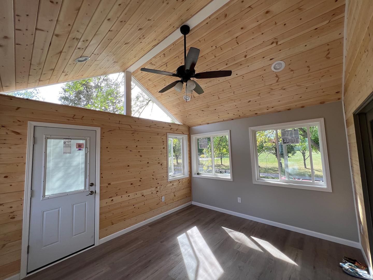 Interior of a small room with wood paneling, windows, and a ceiling fan; sunlight streams in.