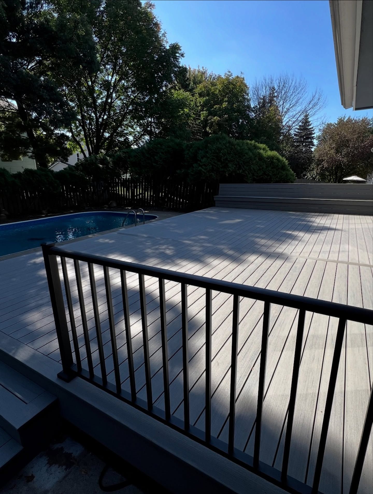 Gray composite deck with black railing and pool in a yard with trees.