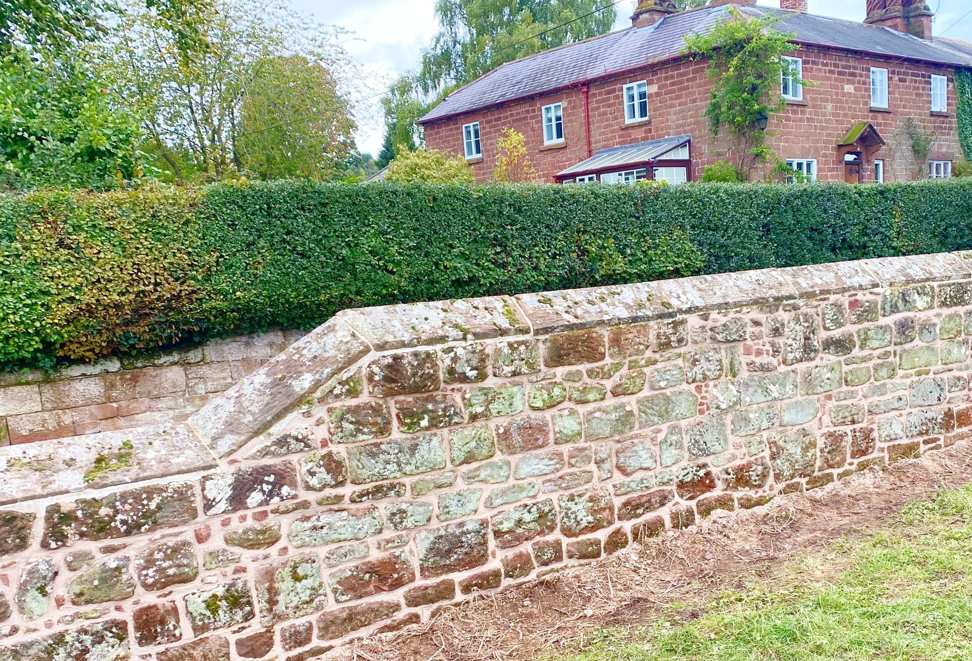 A brick wall with a house in the background and a hedge in the foreground. Stonemasons Cheshire
