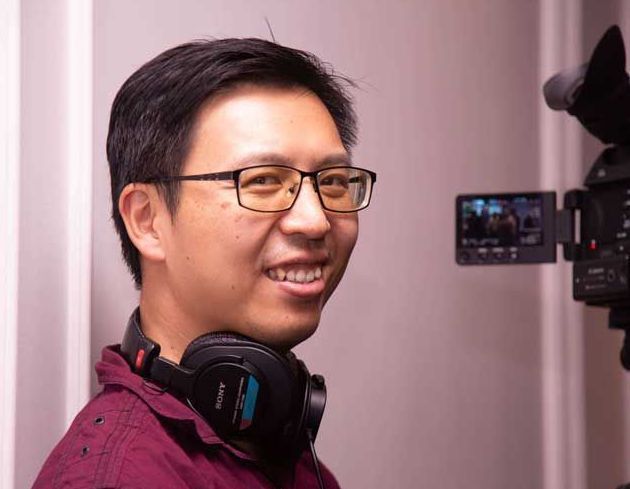 Man with glasses and headphones smiles near a video camera, wearing a red shirt.