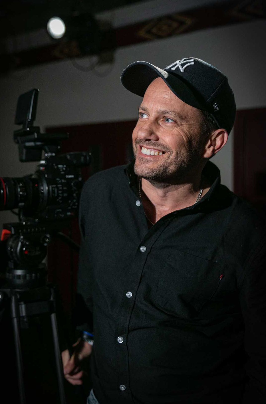 Craig Walsh in a black cap and shirt smiles, stands near a video camera, in a dim interior setting.