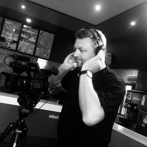 Man adjusting headphones, next to a video camera on a tripod, in a coffee shop setting.