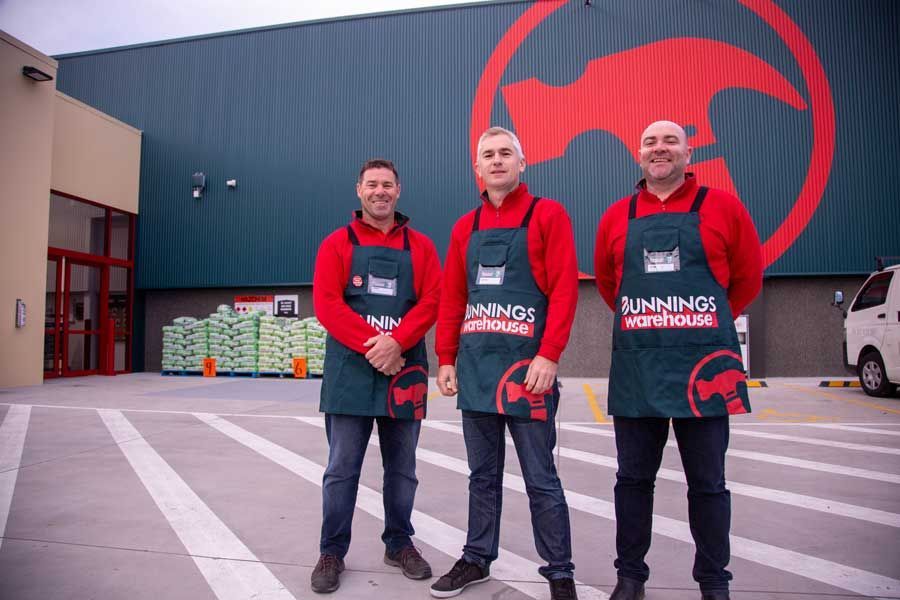 Three people in red shirts and green aprons stand outside a Bunnings Warehouse store.