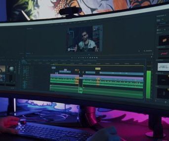Person edits video on a large curved monitor, with keyboard and mouse visible. Neon lighting under the screen.