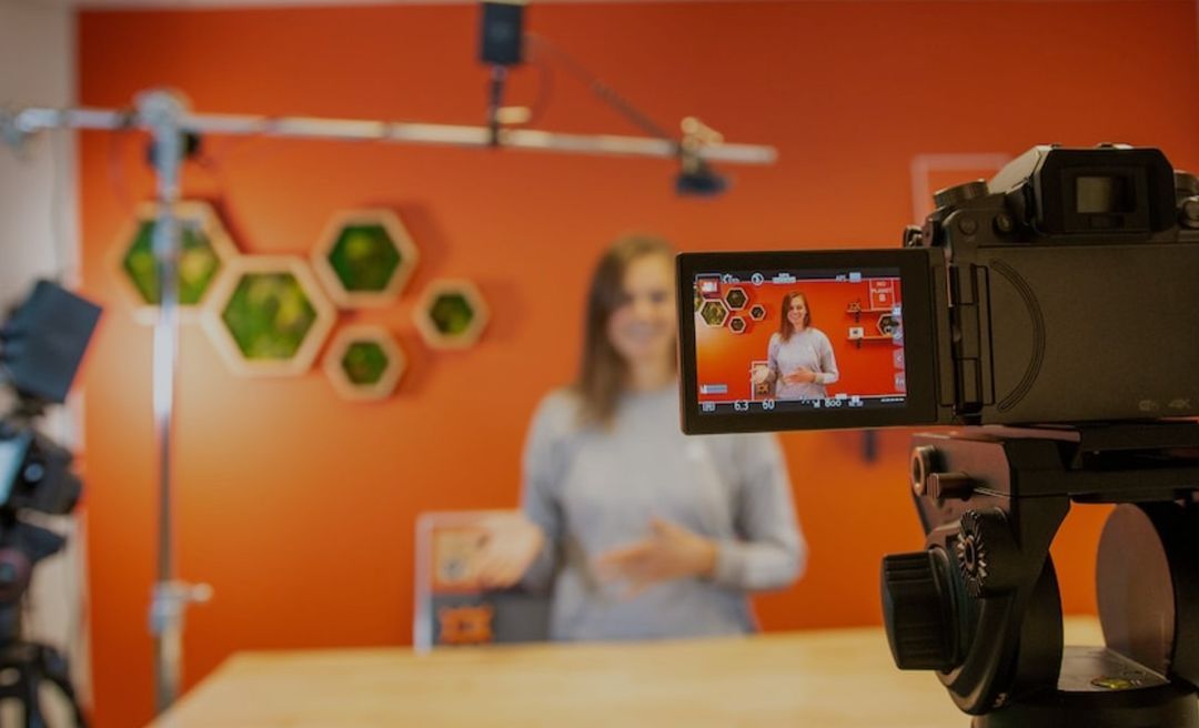 Woman speaking in a video studio with orange wall and camera in focus.