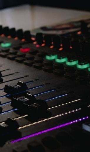 Close-up of a dark audio mixing console with illuminated controls and faders, with green, purple and red lights.