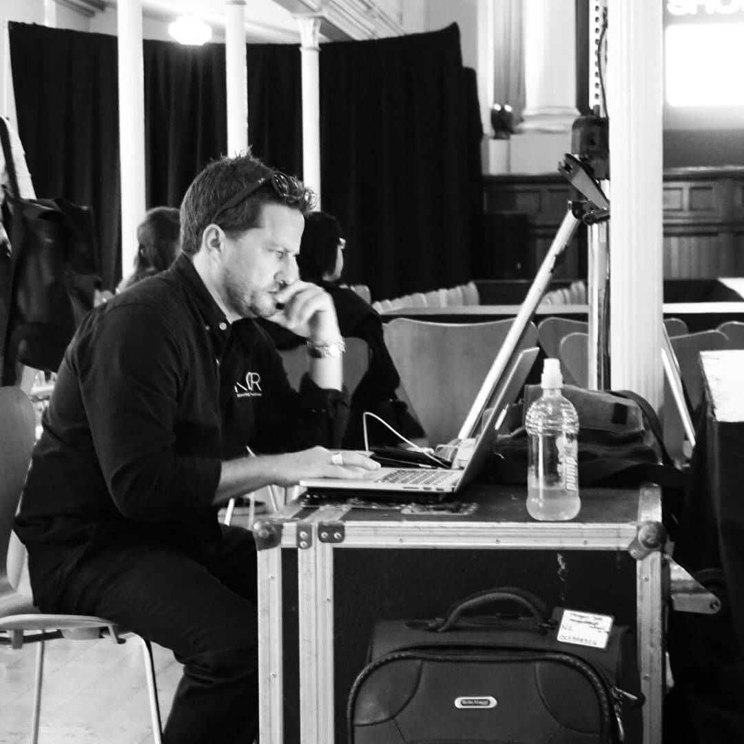 Man working on a laptop, sitting at a table with luggage in an indoor setting. Black and white photo.