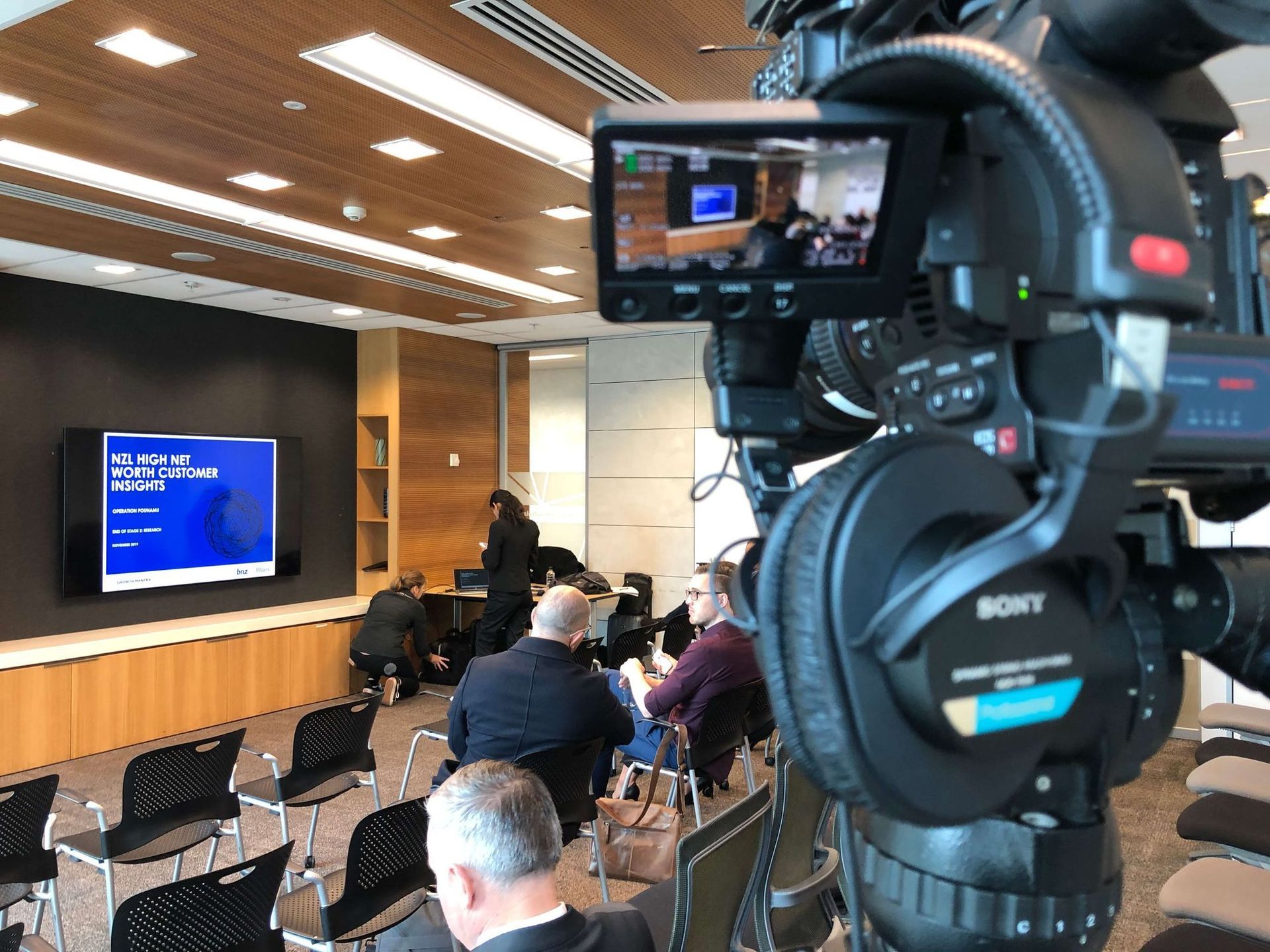 Camera filming a presentation in a modern room with a screen and seated audience.