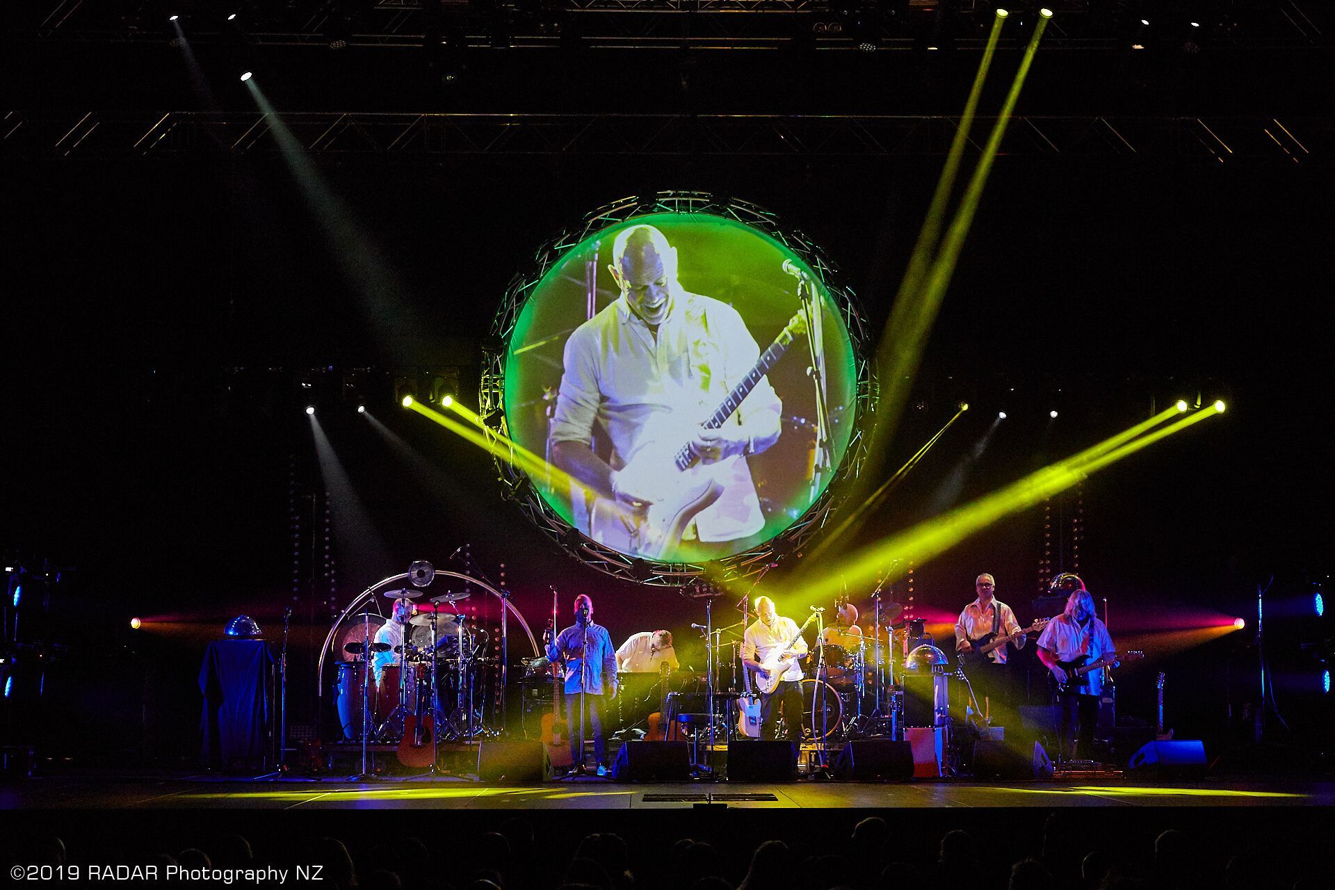 Pink Floyd Experience Band performing on stage with a large screen showing a musician. Bright yellow spotlights.