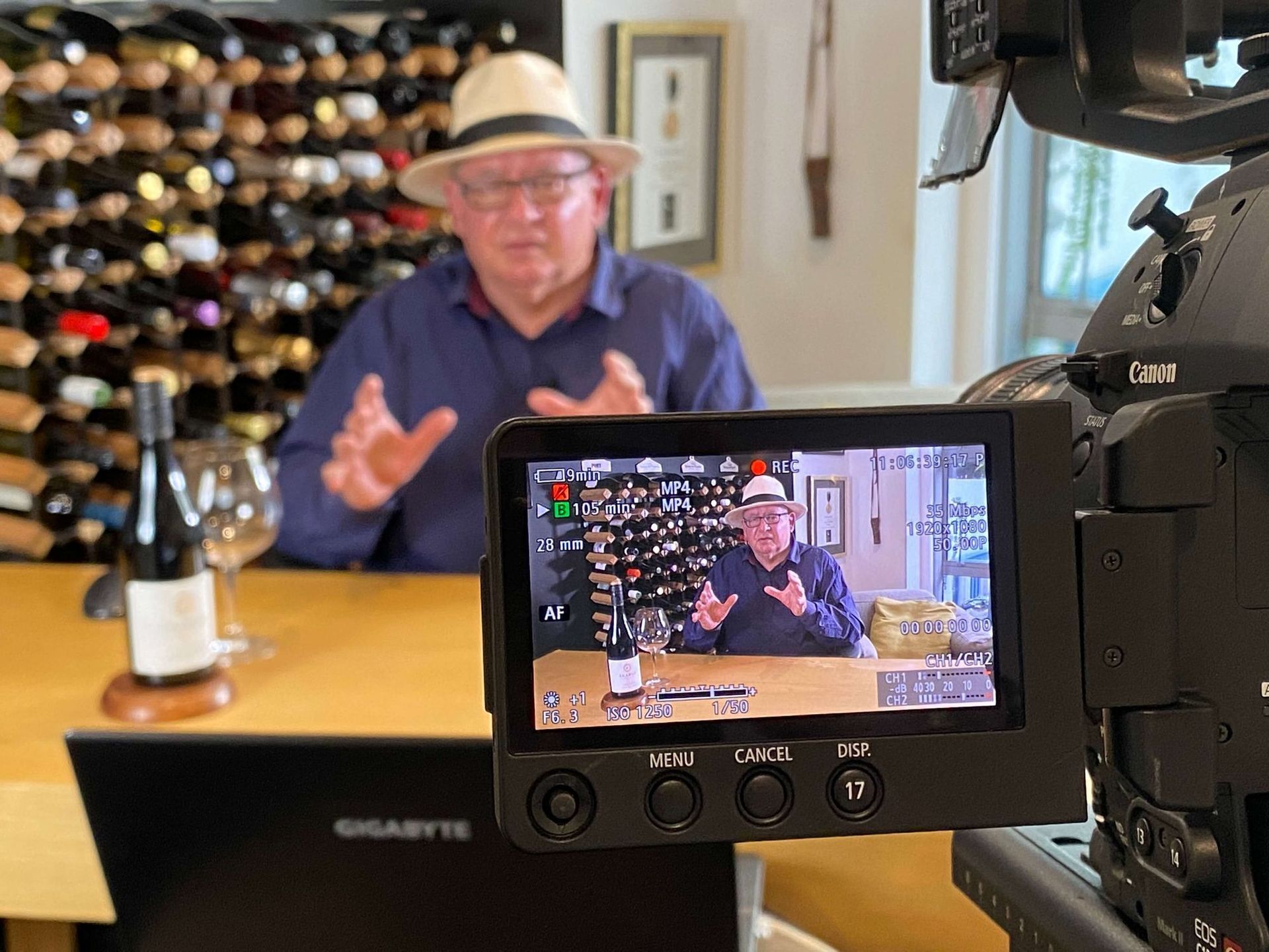 Man in hat gestures during a video recording in a wine cellar. A monitor shows the same scene.