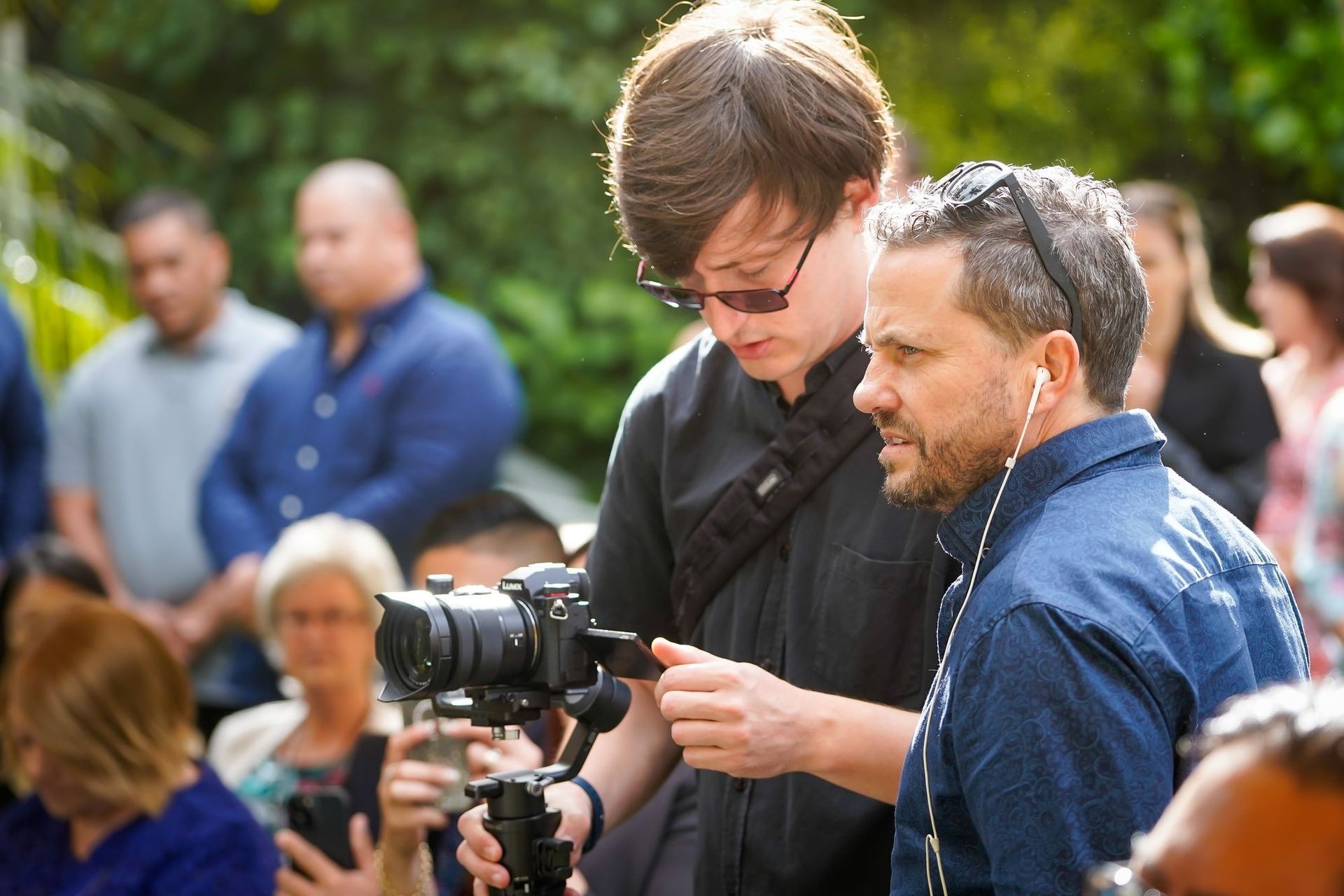 Two men filming outdoors with a camera on a tripod. Other people are in the background, mostly blurred.