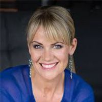 Blonde woman with blue top smiles, wearing dangling earrings, indoors.