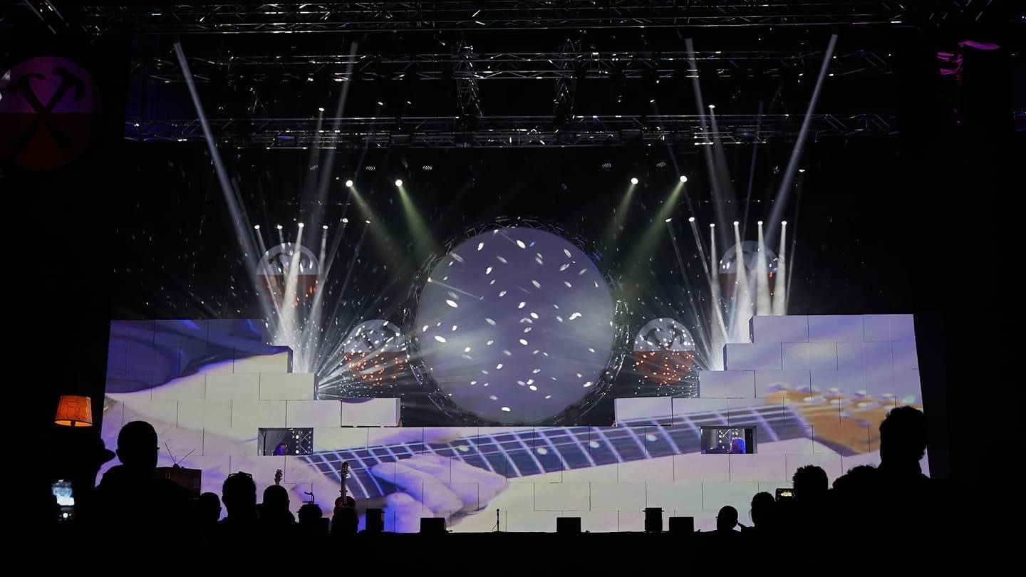 Concert stage with large screen displaying a guitar neck. A central sphere and side structures glow with lighting.
