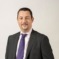 Man in dark suit, purple tie, smiling, indoors, against a white background.