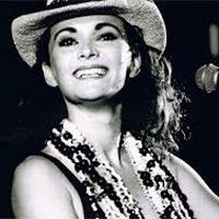 When the Cats Away singer wearing a cowboy hat and beaded necklace smiles at a microphone in a dark setting.