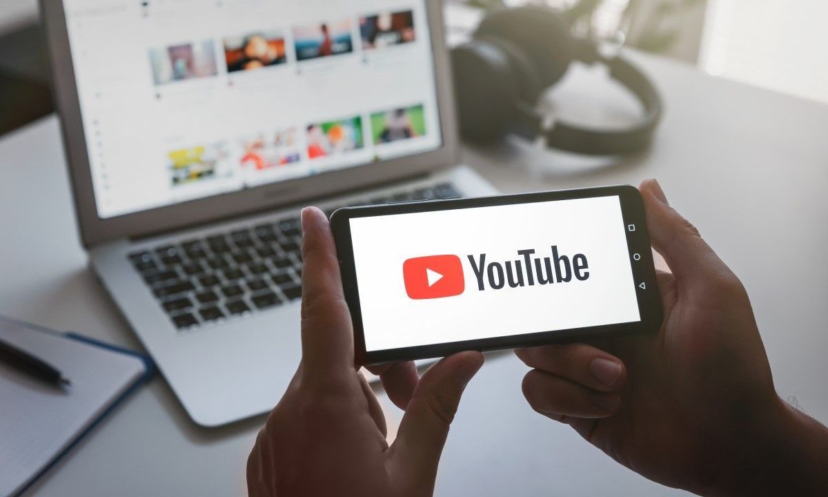 Person holding phone displaying YouTube logo, laptop and headphones in background.