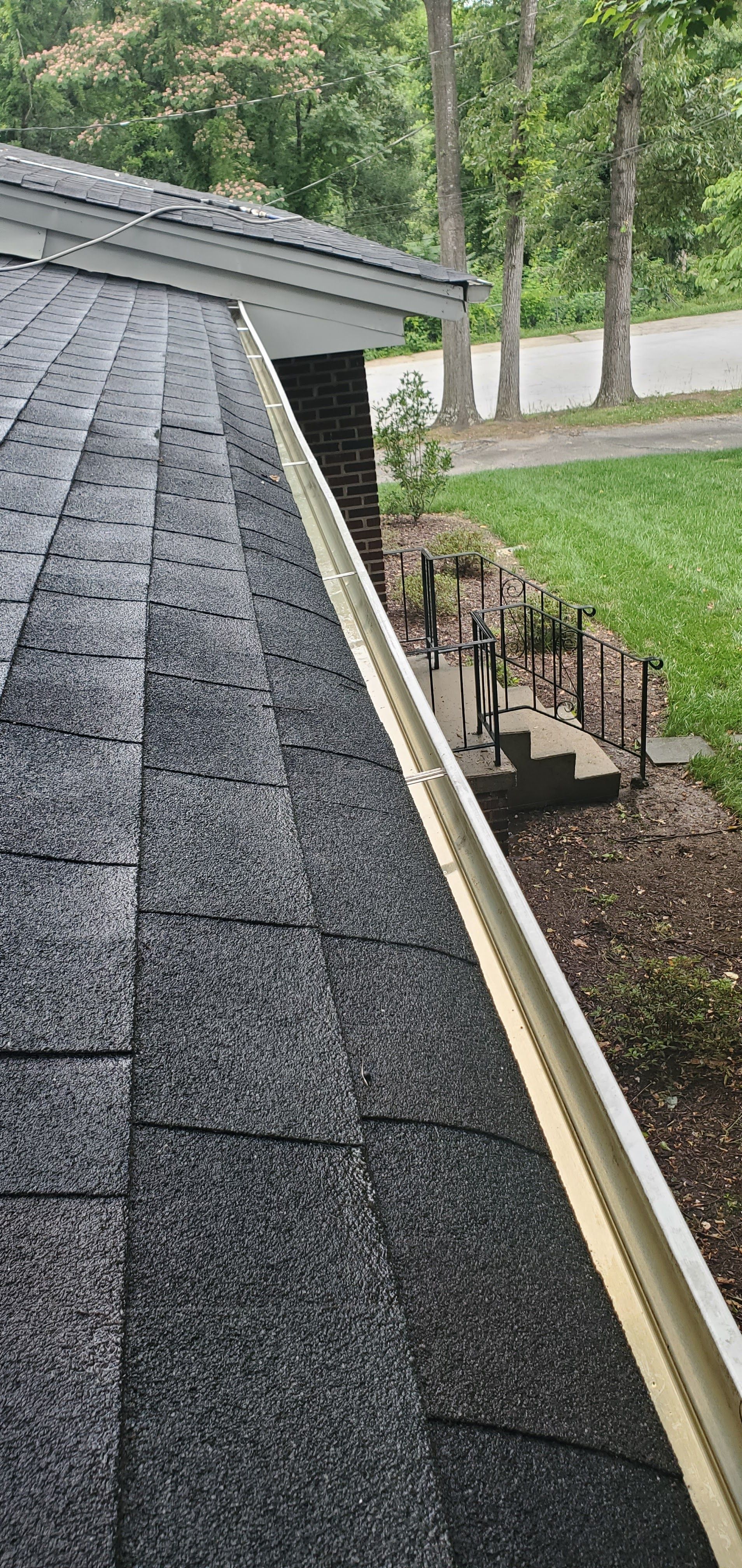 Roofline with dark shingles and gutter beside a landscaped yard with trees and a road in the background