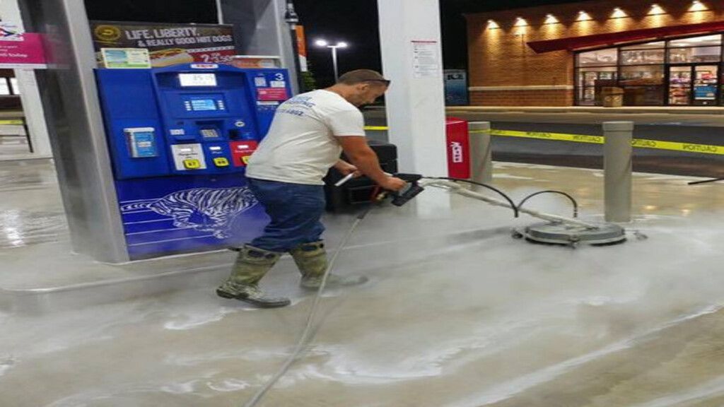 Worker pressure-washing a gas station pump area at night, with water spraying across the wet ground.