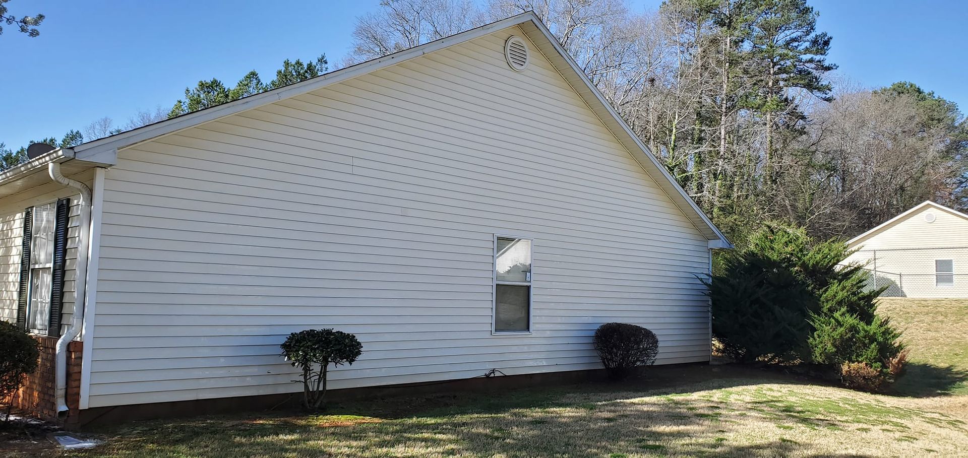 White vinyl house exterior with a steep roof, small arched window, and shrubs on a sunny lawn.