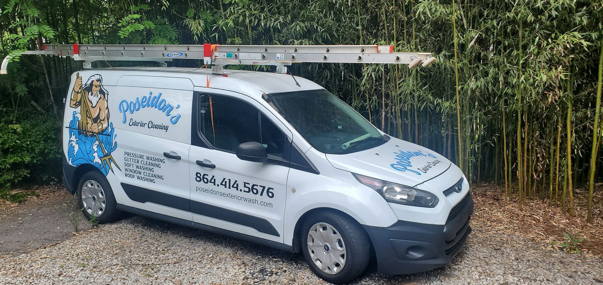 White service van with blue graphics and ladder on top parked on a gravel roadside near greenery