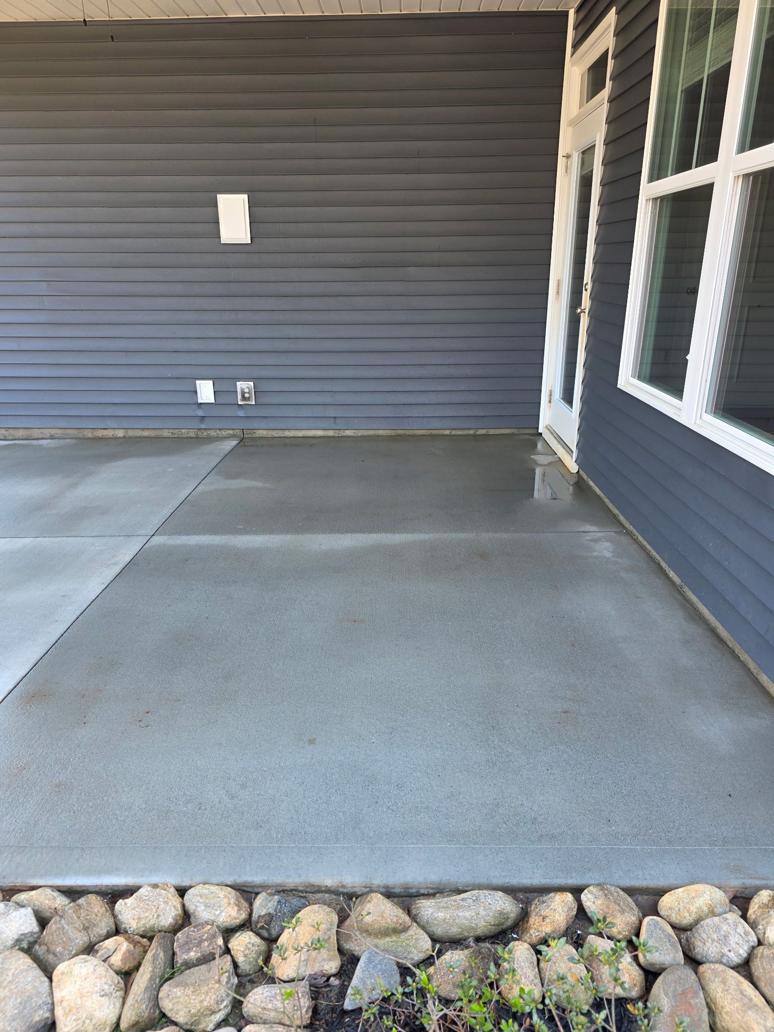 Empty concrete front porch with gray siding and windows, bordered by landscaping rocks