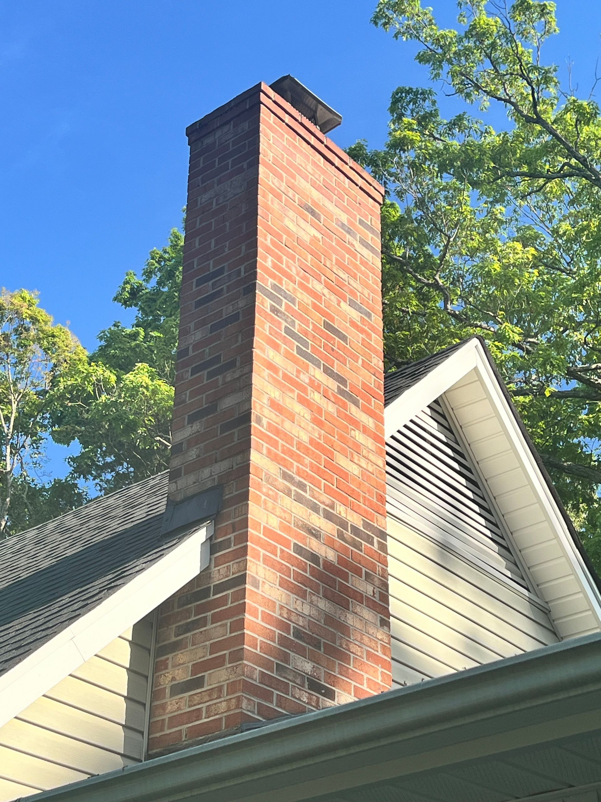 Brick chimney rising above a house roof against a blue sky and green tree branches