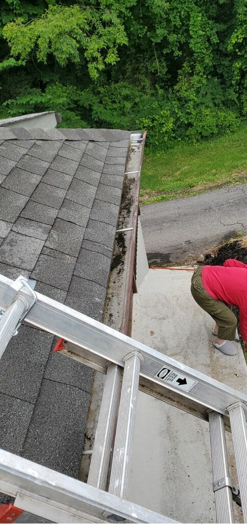 Person in red shirt working on a roof beside a ladder, with trees and a road below.