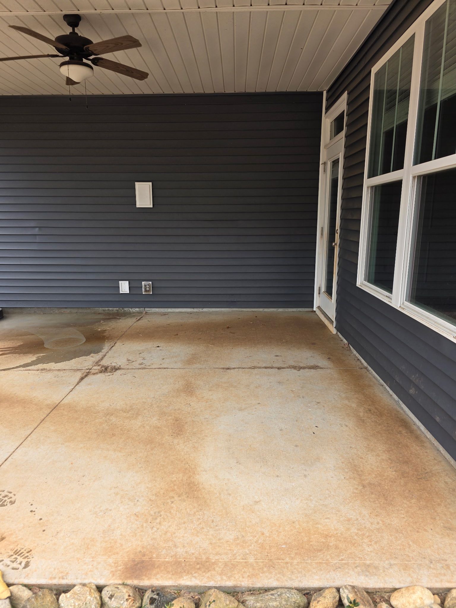 Covered concrete patio with dark siding, ceiling fan, and windows on the right.