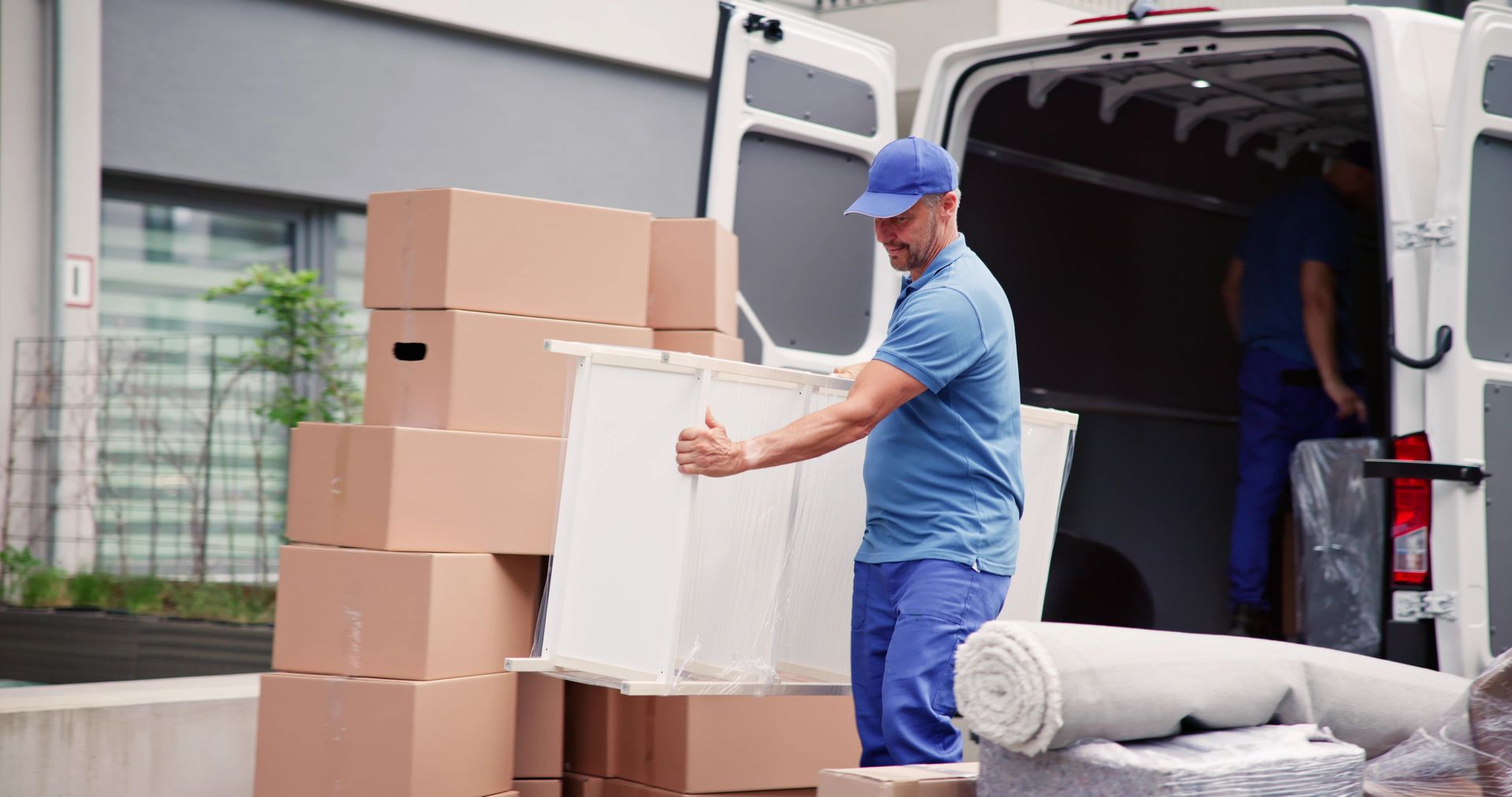 Movers loading a white panel onto a van, boxes and rolled carpet nearby.
