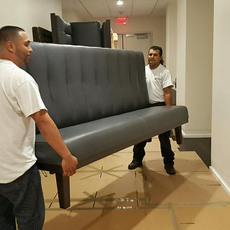 Two people in a hallway carrying a gray futon, likely moving furniture.