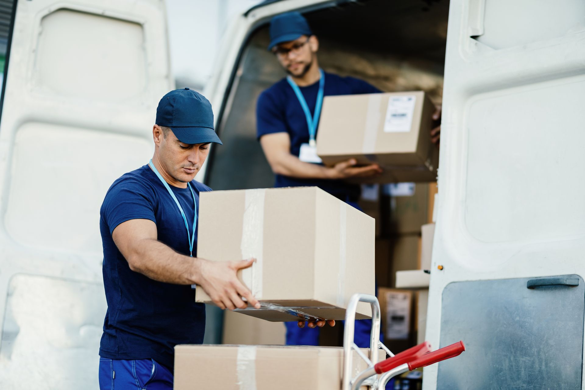 Two delivery workers loading cardboard boxes from a van onto a hand truck.