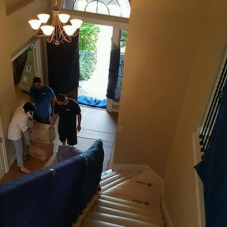 Movers carrying boxes and furniture into a house with a staircase and a chandelier.