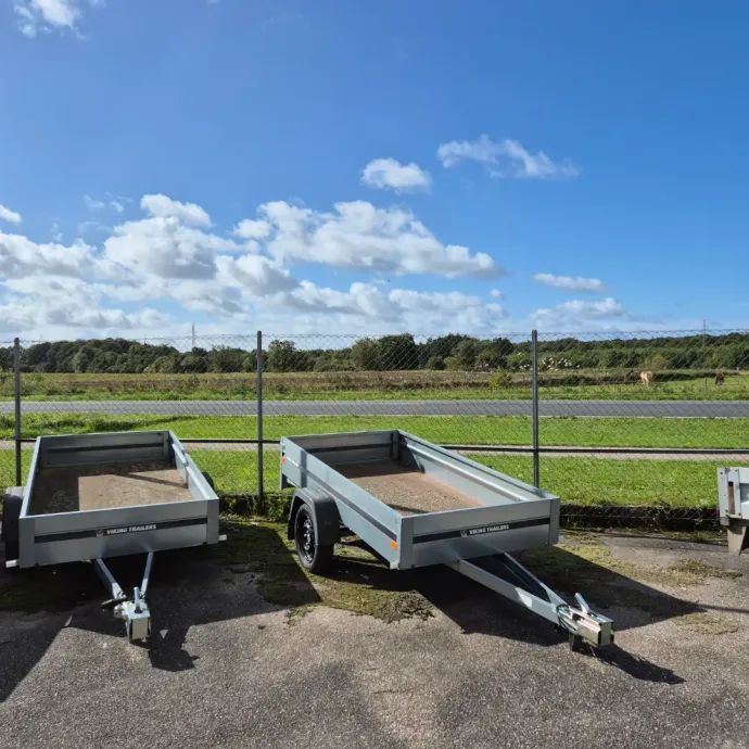 Tre grå utility trailere parkeret på asfalt med grønt græs og et hegn foran en vej og træer, blå himmel.