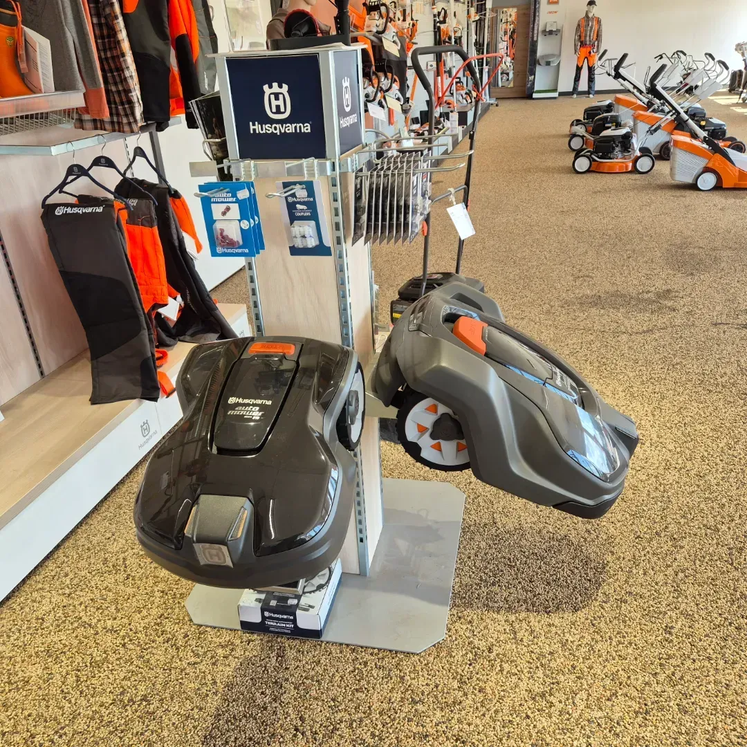 To robotplæneklippere udstillet i en butik, med tilbehør og andre plæneklippere synlige i baggrunden.