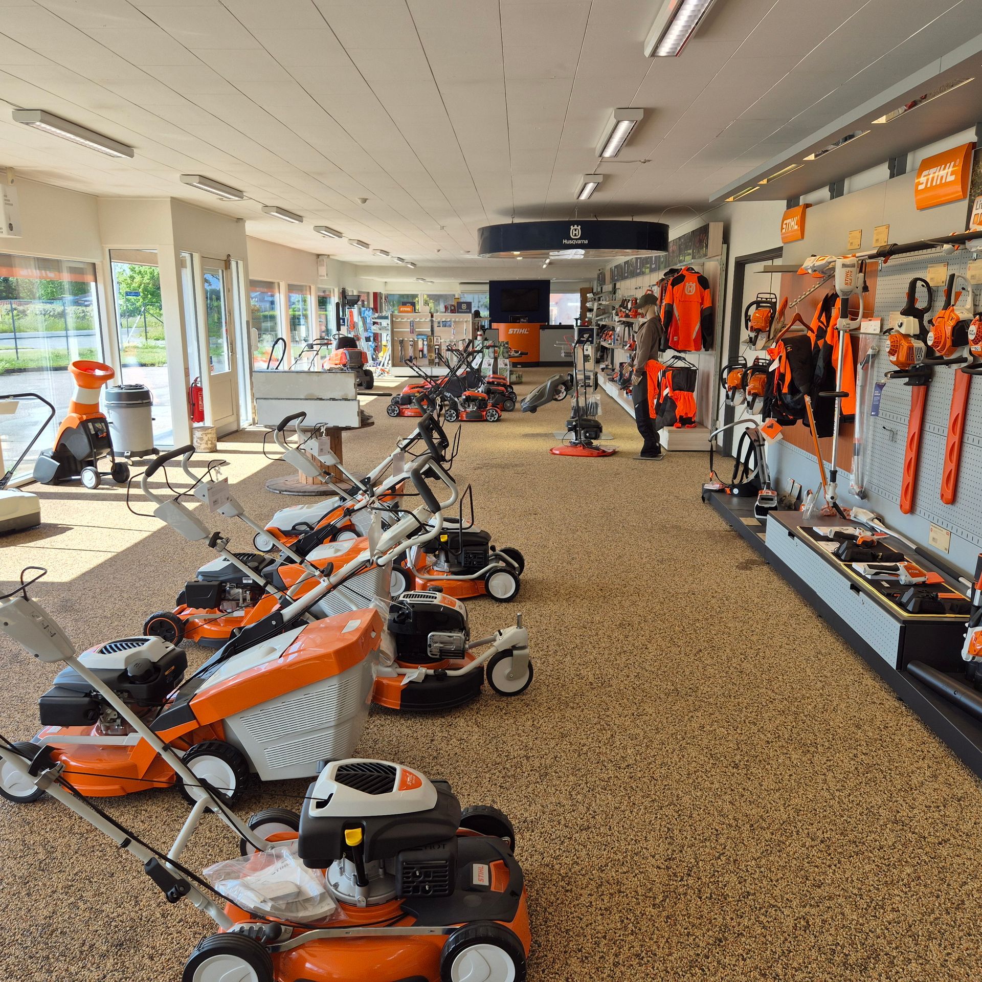 Inde i en butik med plæneklippere og værktøj til salg, hvor der sælges forskellige orange og sorte plæneklippere.