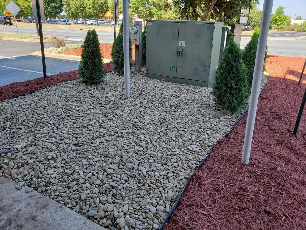 Small landscaped utility area with white gravel, red mulch, shrubs, and gray electrical boxes beside a walkway.