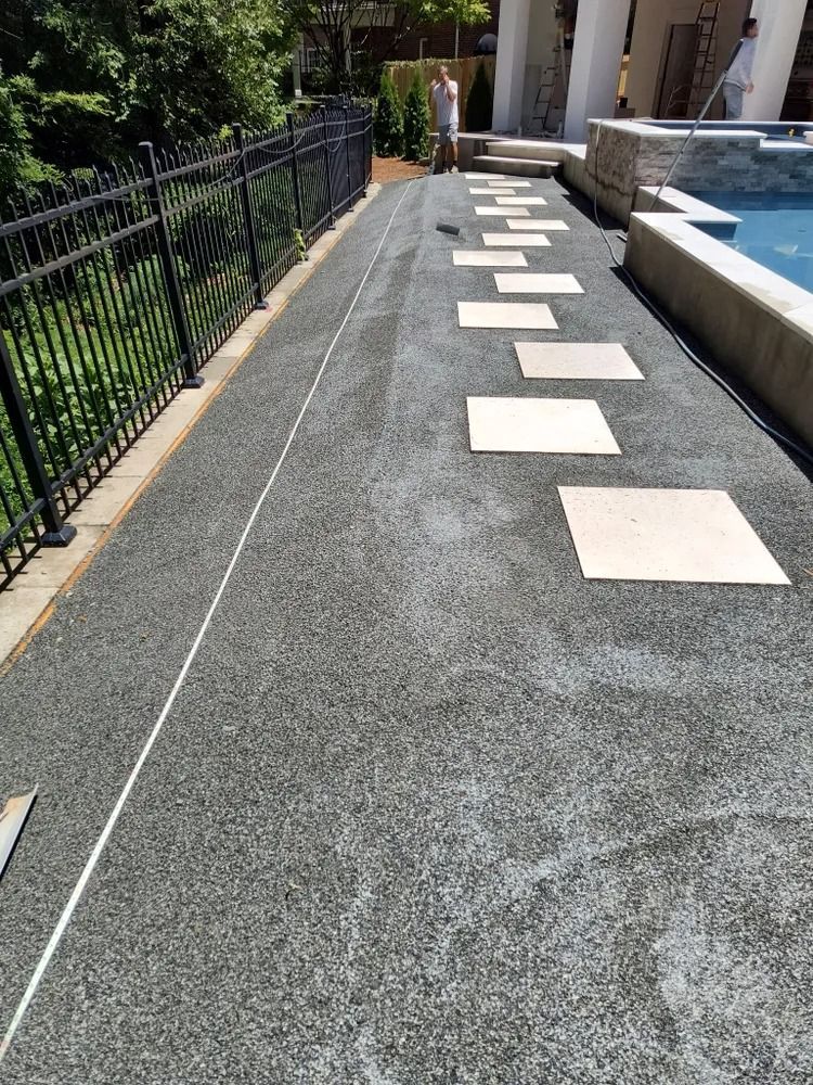 Outdoor pathway with square stepping stones beside a black fence and landscaped pool edge.
