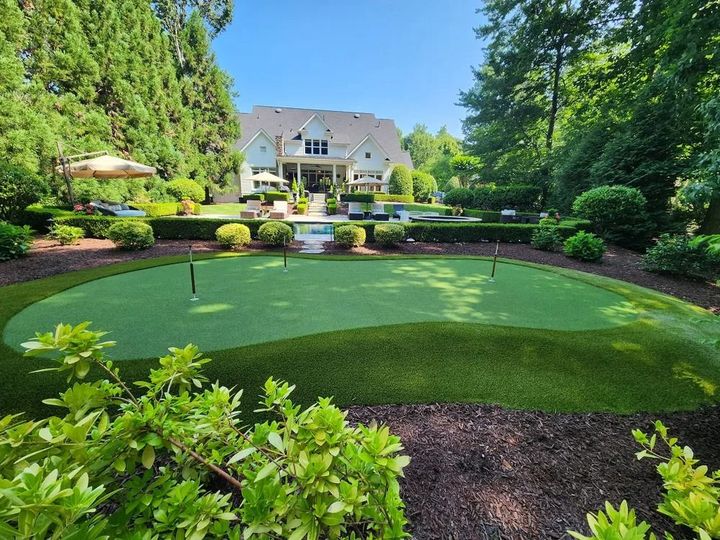 Lush backyard putting green with flagsticks, surrounded by manicured garden and a large house in the background