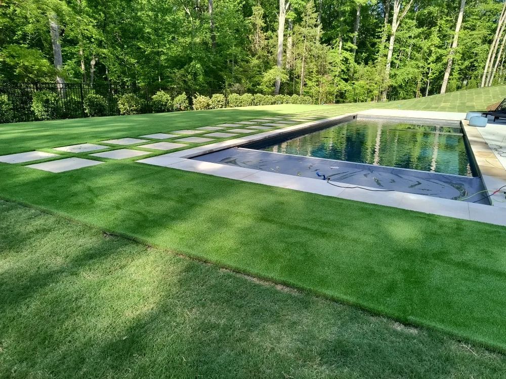 Rectangular backyard pool with stone patio, green lawn, and trees in the background