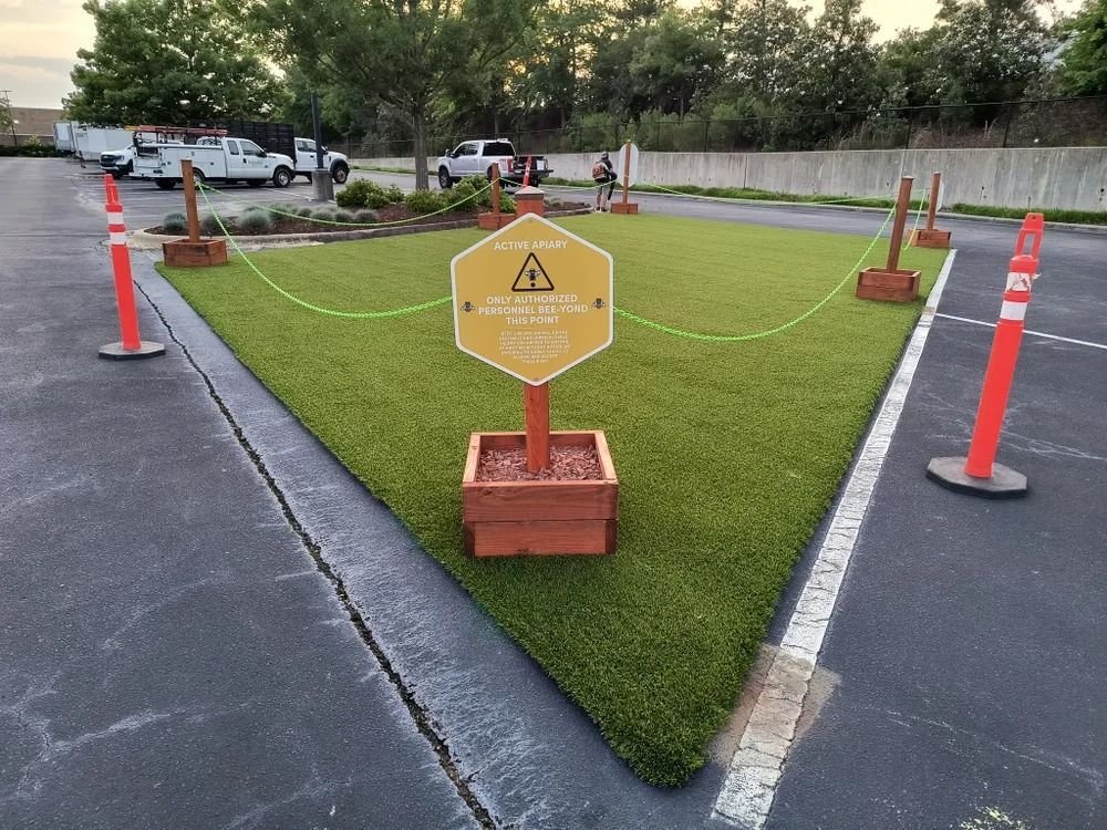 Yellow caution sign in a small cordoned-off grassy area, bordered by orange cones and red rope near a sidewalk.