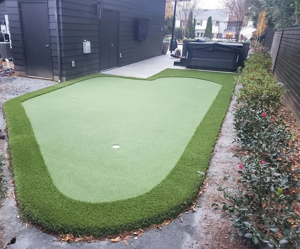 Small backyard putting green with green artificial turf bordered by a darker fringe, beside a modern house and fence