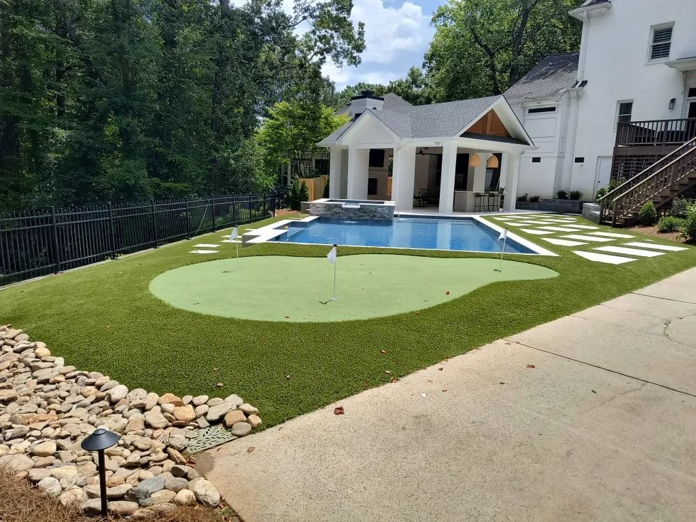 Backyard pool with green artificial turf, white patio stones, and a white house in the background