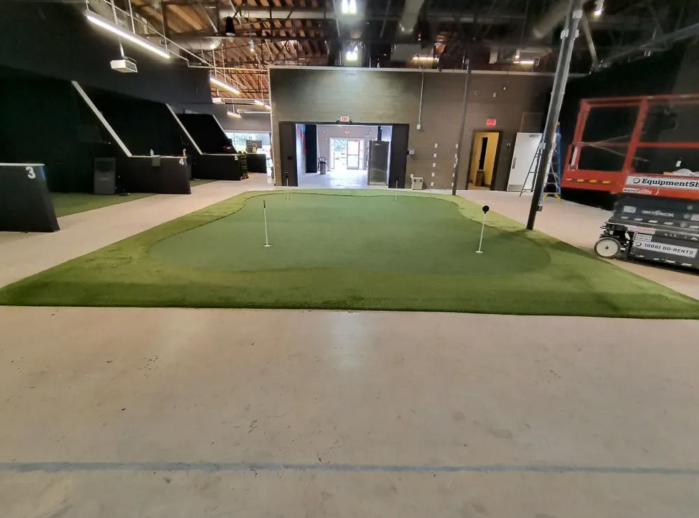 Indoor golf putting green with a flagstick in a warehouse-like space, surrounded by concrete floor and equipment booths