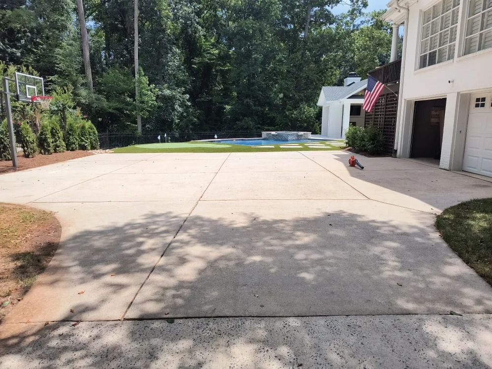 Concrete backyard driveway with a basketball hoop, pool, and house garage in the background