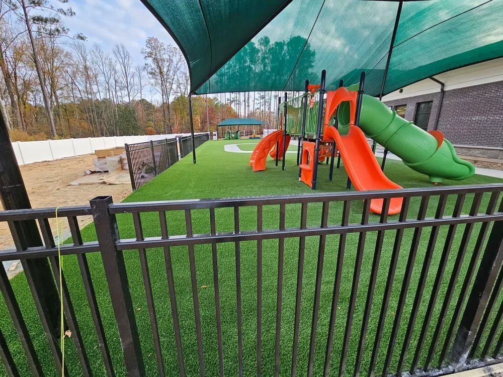 Empty playground with orange and green slides under shade netting, viewed from behind a black fence