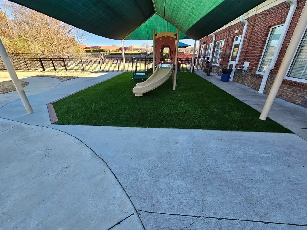 Covered playground with a tan slide beside a brick building and green shade sails.